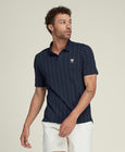 A man modeling a navy blue short-sleeve polo shirt with vertical stripes, paired with off-white shorts, white socks, and white sneakers, against a solid light beige studio background.